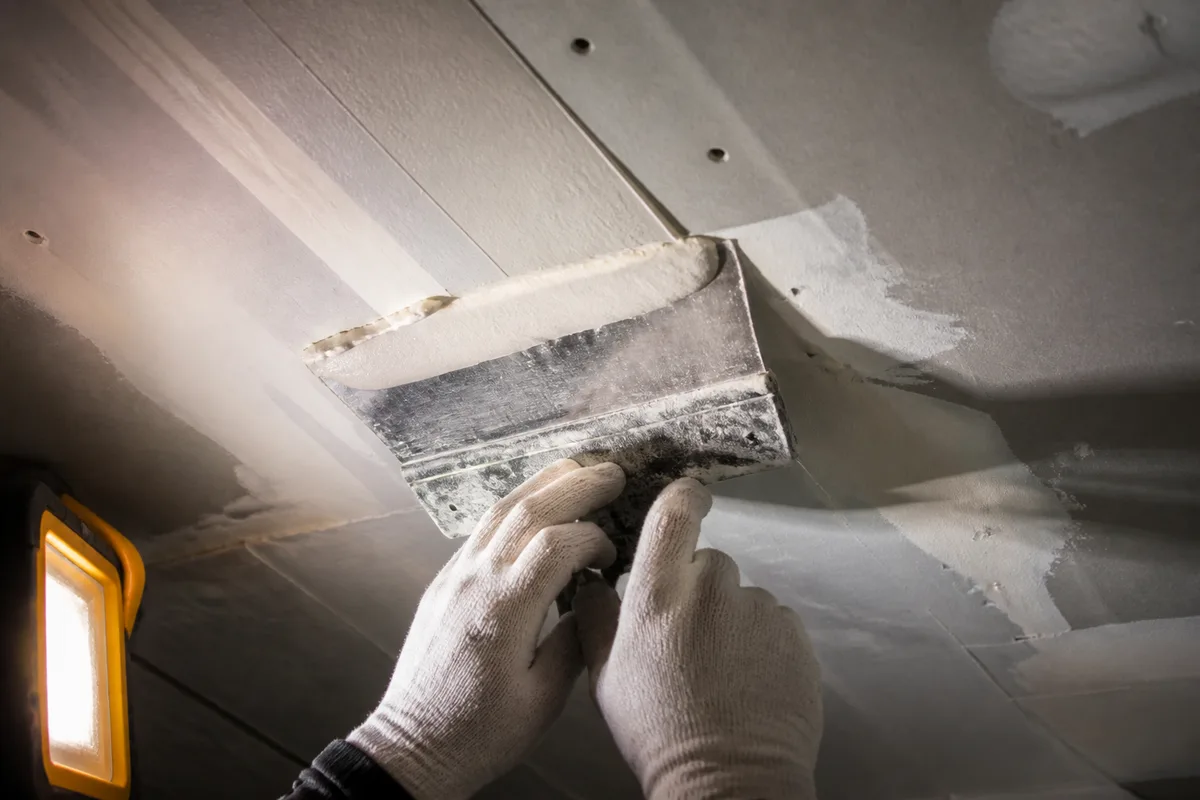 Plafond en cours de préparation, mains gantées appliquant un enduit de lissage à la spatule large